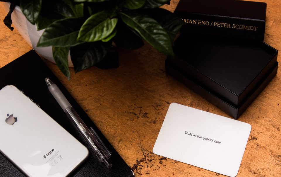 A white card that says "Trust in the you of now" sits on a brown table next to to black boxes, an iphone, and a plant. The card is from a pack designed to help people get inspiration, and possible grow your business.