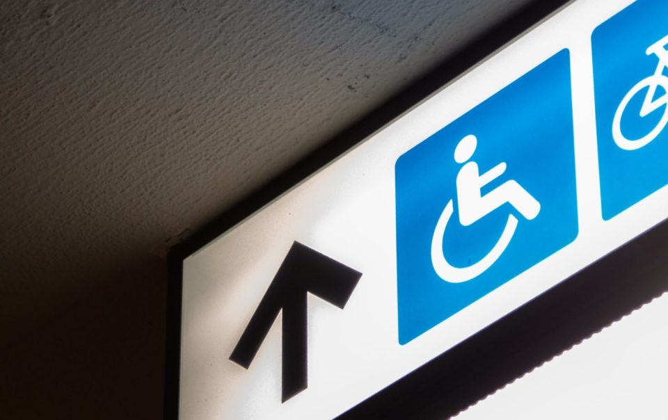 A fluorescent white sign on which there is a blue image of a person sitting in a wheelchair. Next to the image is a black arrow pointing forward. The image as a whole accompanies an article about increasing accessibility in one's business.