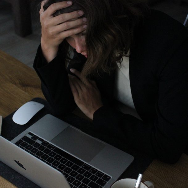 A woman sitting at a desk in front of a laptop with her head in her hands. It accompanies an article about hustle culture.